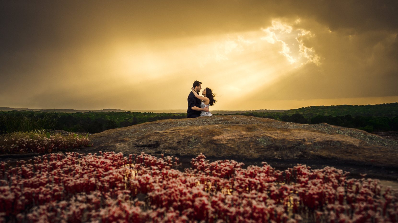 couple hugging mountain top with sun piercing through storm clouds red flower diamorpha blooming