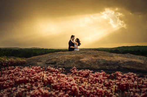 couple hugging mountain top with sun piercing through storm clouds red flower diamorpha blooming