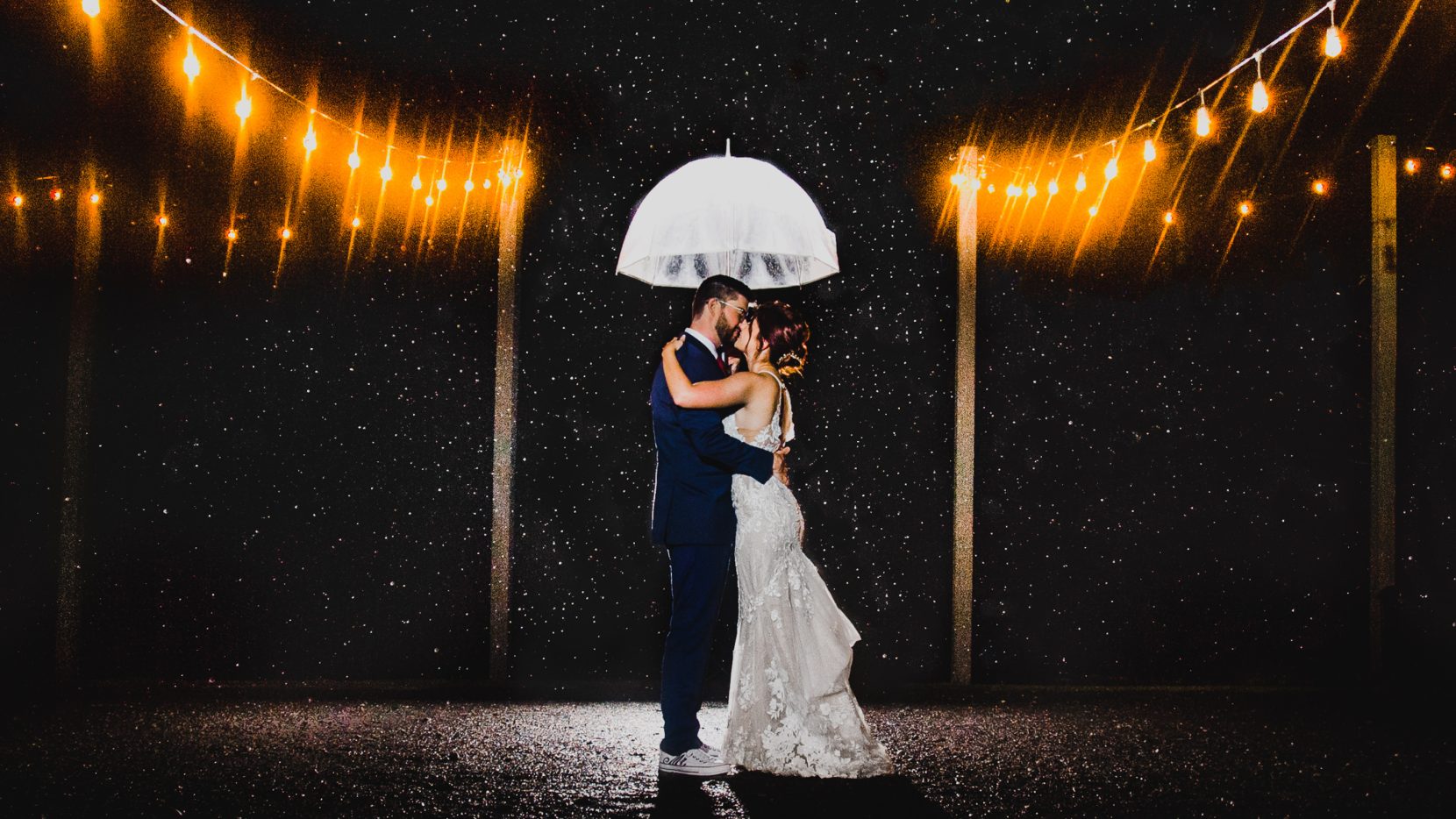 bride groom kissing under clear umbrella rain with cafe lights